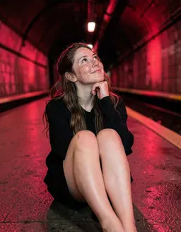 freckledgirl, a 22‑year‑old caucasian girl with a freckled complexion, long wavy brown hair and soft facial features, sits on the cold subway floor with her knees drawn up, chin lifted and eyes gazing upward, a subtle smile playing on her lips, dressed in a loose black long‑sleeved top. the scene glows with ruby‑red light filtering from the tunnel walls, casting warm crimson reflections on the damp concrete and highlighting the gentle curve of her pose, while the surrounding subway tracks disappear into the glowing darkness.