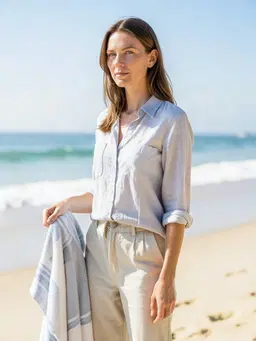 Replace the hybrid portrait with a British adult woman featuring ash‑brown hair and green eyes, dressed in relaxed casual clothing (e.g., a light‑washed shirt and soft trousers) and positioned on a sunny beach. Pose her standing with one hand lightly holding a beach towel, the other relaxed at her side, against a backdrop of gentle waves and a clear sky; use bright, natural daylight from a slightly side‑angle to create soft shadows and a warm, inviting atmosphere. Preserve the subject’s human silhouette, facial structure, and overall composition while swapping the red hair, pale complexion, and mosaic‑textured skin for the specified ash‑brown hair, green eyes, and natural skin tone. Do not alter the subject’s gender, age, or basic facial proportions.